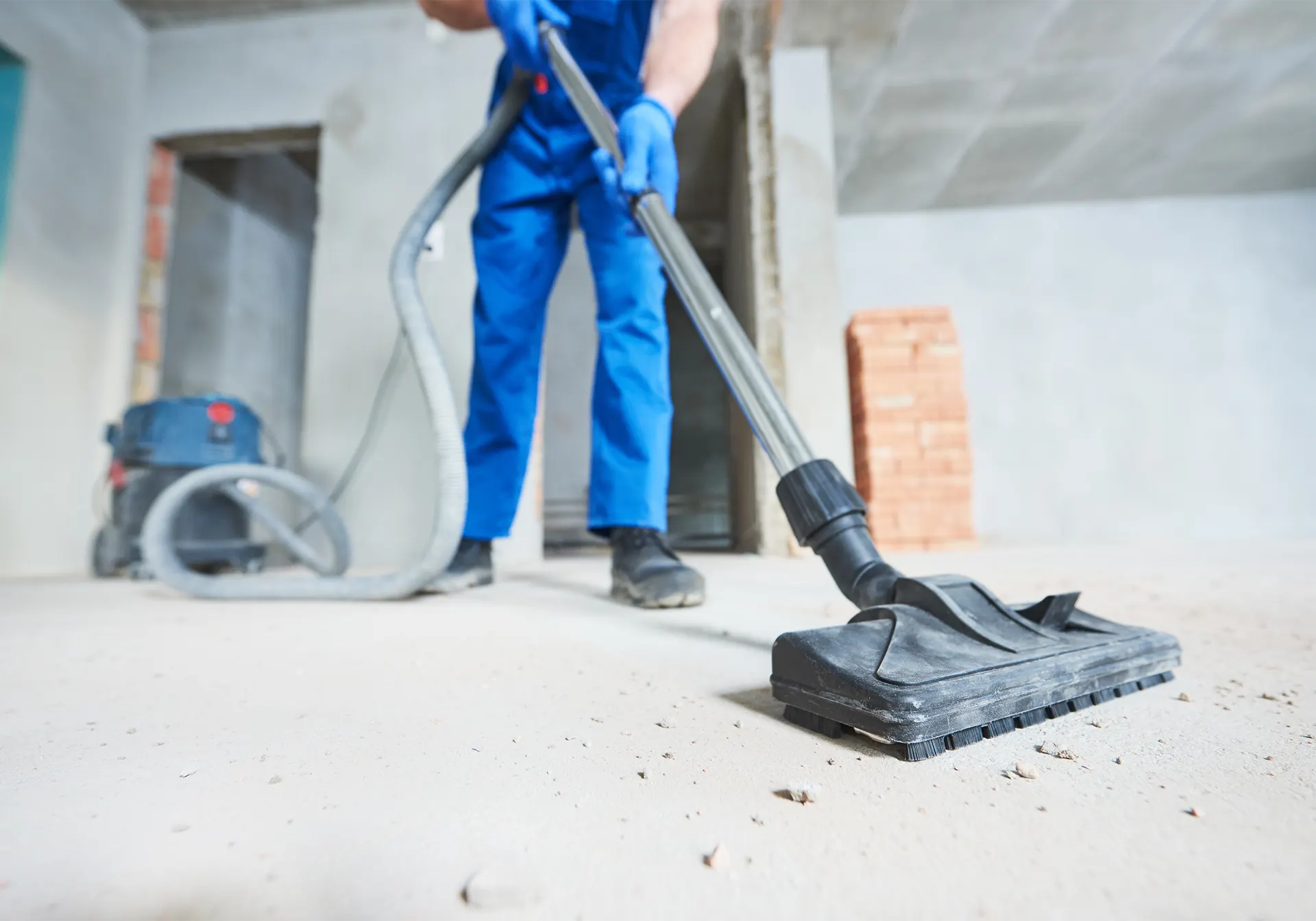 Person in blue overalls vacuuming a floor near a stack of bricks and a dustpan